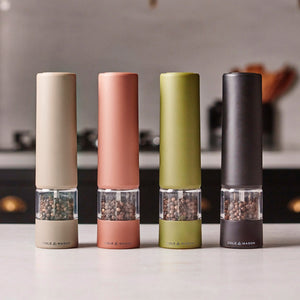 Four Cole & Mason Brixton Electric Salt and Pepper Mills—including the Olive Green GS—stand in a row on a kitchen counter in beige, pink, green, and black, each filled with peppercorns. Designed with durable ceramic grinders for smooth, one-handed operation, these mills add both style and convenience to any kitchen. A softly blurred kitchen appears in the background.