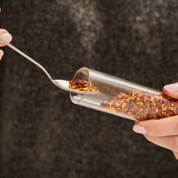 A person holds the Cole & Mason Shadwell Slim-Mount Spice Storage filled with red chili flakes, scooping some out with a spoon against a dark background.