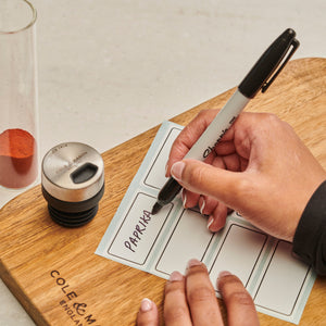 A hand writes PAPRIKA on a spice label with a black marker beside a jar and lid—key parts of the Cole & Mason Shadwell Drawer-Fit Spice Storage, an ideal kitchen storage solution.