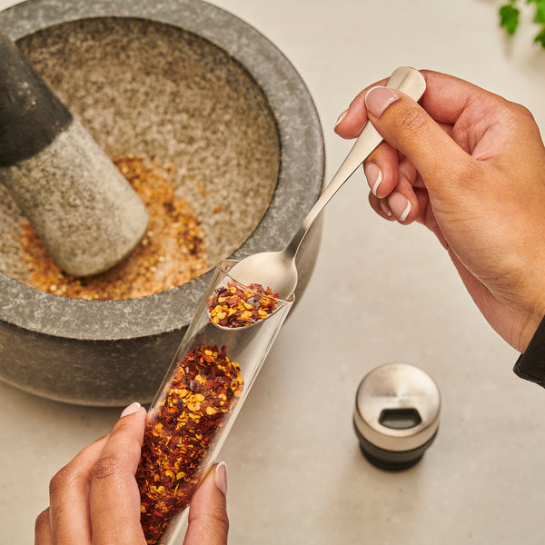 A person spoons dried chili flakes from the Cole & Mason Shadwell Drawer-Fit Spice Storage into a stone mortar, with a pestle and spice lid nearby on a light countertop.