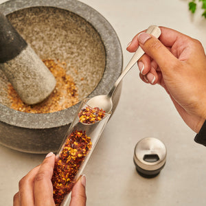 A person scoops dried chili flakes from a Shadwell Countertop Spice Storage by Cole & Mason, beside a stone mortar and pestle—ideal for keeping your spices organized on the countertop.