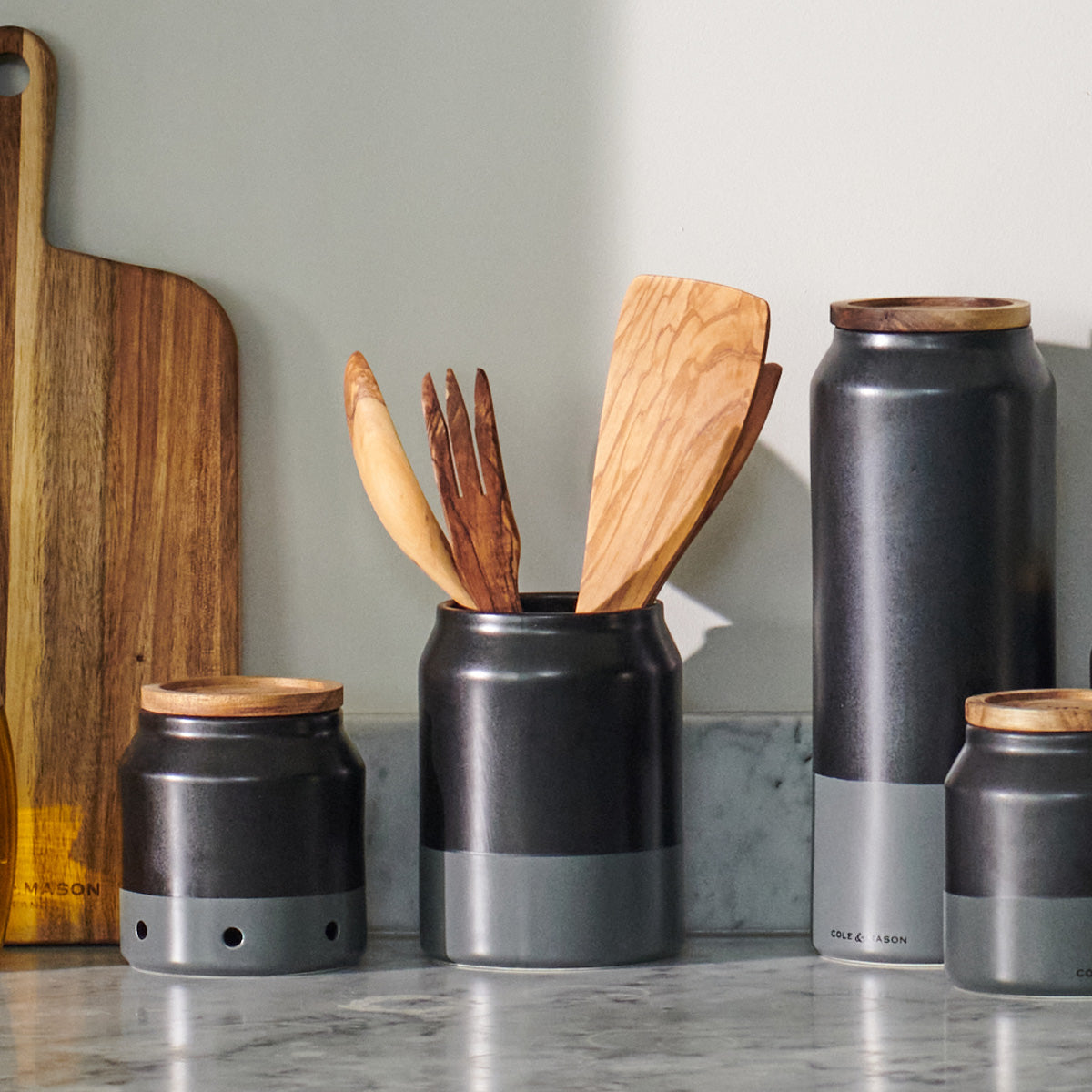 A set of kitchen canisters and wooden utensils are arranged on a marble countertop next to a wooden cutting board, with sunlight casting soft shadows.