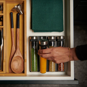 A tidy kitchen drawer with wooden utensils, green cloths, and a hand reaching for an airtight glass jar among spices stored in the Cole & Mason Shadwell Drawer-Fit Spice Storage organizer.