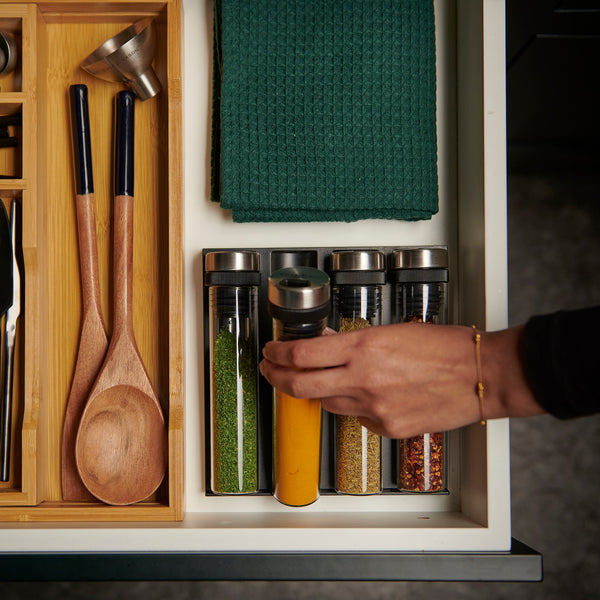 A tidy kitchen drawer with wooden utensils, green cloths, and a hand reaching for an airtight glass jar among spices stored in the Cole & Mason Shadwell Drawer-Fit Spice Storage organizer.