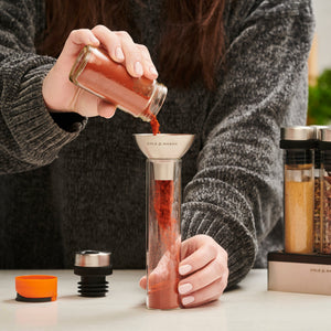 A person pours red spice from an airtight glass jar into the Shadwell Drawer-Fit Spice Storage by Cole & Mason using a metal funnel, with spice grinders and kitchen items on the countertop nearby.
