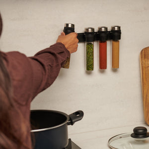 Wearing a brown shirt, a person reaches for the wall-mounted Cole & Mason Shadwell Slim-Mount Spice Storage, featuring four airtight glass containers above a kitchen counter with a pot and wooden cutting board.