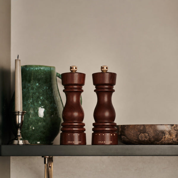 A green ceramic pitcher, a white candle, the Cole & Mason London Walnut Salt or Pepper Mill, an adjustable salt grinder, and a brown bowl are arranged neatly on a gray shelf against a beige wall.