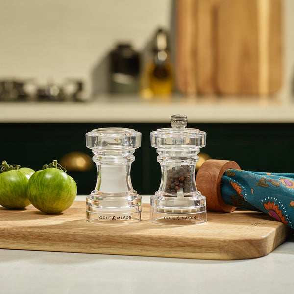 A set of Cole & Mason Acrylic Salt Shaker & Pepper Mill, 104 mm, with adjustable grinding mechanisms for personalized seasoning, rests on a wooden cutting board beside green tomatoes and a blue cloth in a warm kitchen atmosphere.