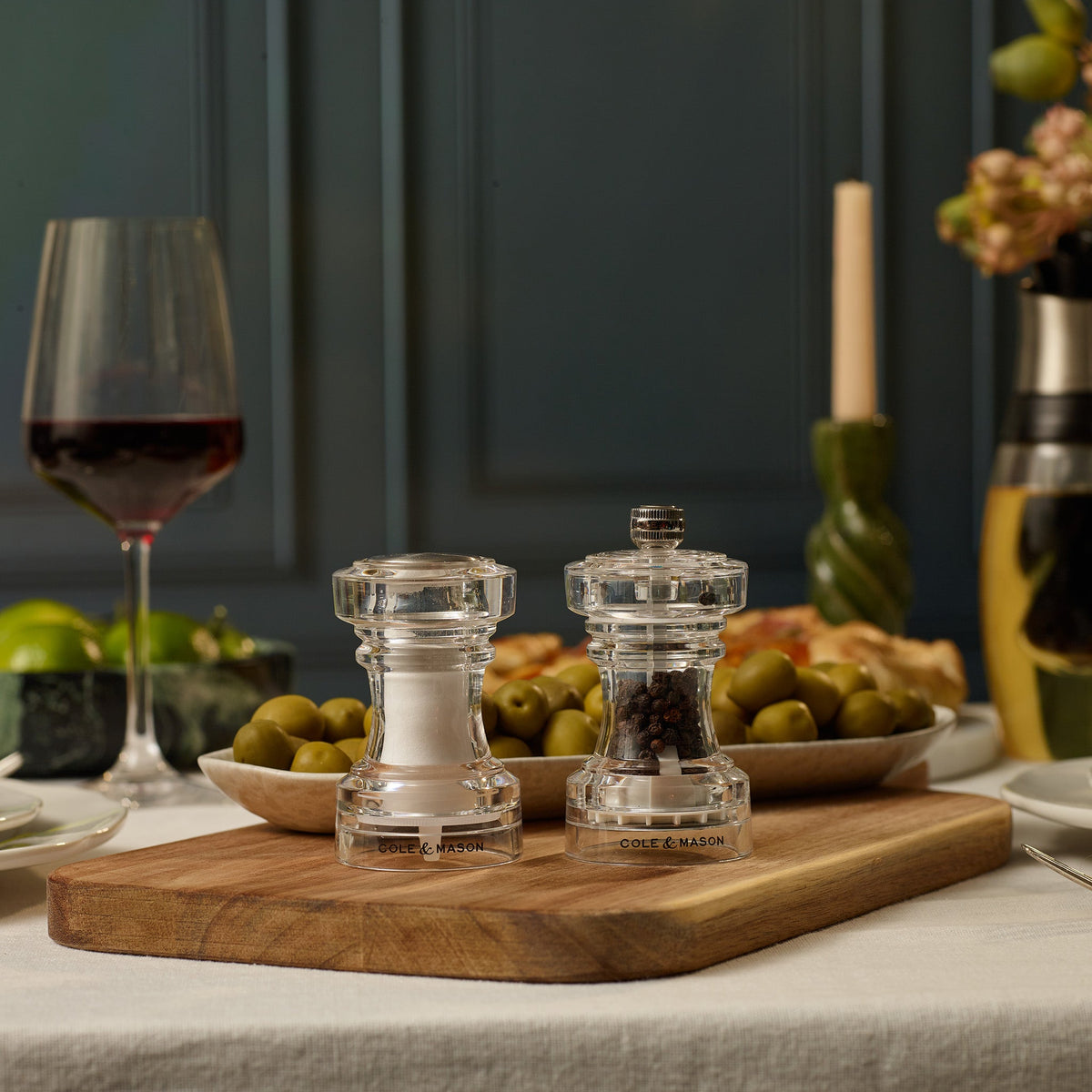 Two Cole & Mason Acrylic Salt Shaker & Pepper Mill Set, 104 mm, with adjustable grinder settings rest on a wooden board amidst bowls of green olives, a glass of red wine, and a partially visible candle, all arranged on a white tablecloth.