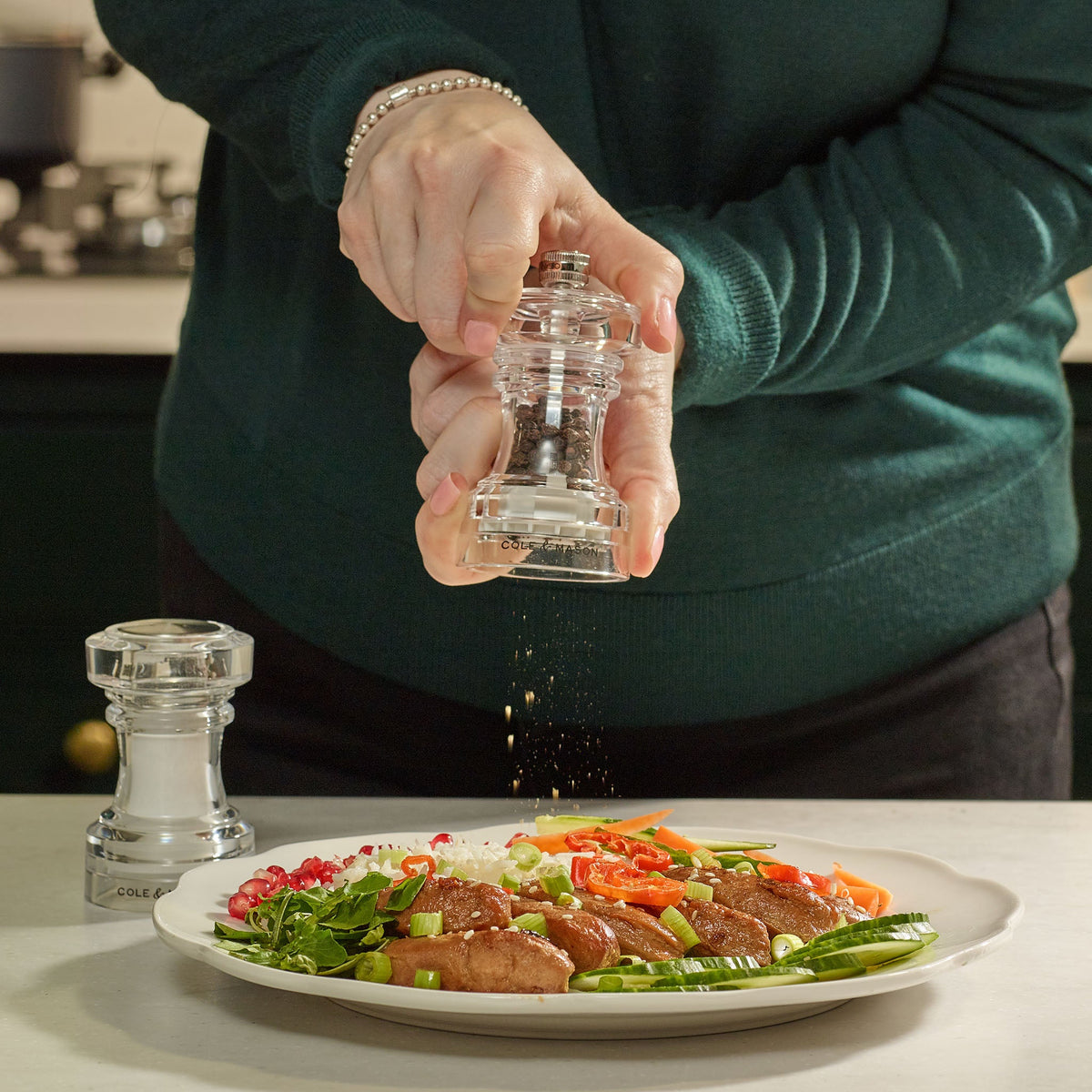 A person in a green sweater grinds pepper onto a plate of food with vegetables and meat, using the Cole & Mason Acrylic Pepper Mill from the 104 mm Salt Shaker & Pepper Mill Set. Another grinder from the set rests on the table nearby, next to the matching acrylic salt shaker.
