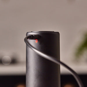 A Cole & Mason Brixton Electric Mill Matt Black GS, featuring a sleek cylindrical design with a power cord plugged in and a small red indicator light glowing near the port, is showcased against a blurred background that accentuates its convenient one-hand operation in the foreground.