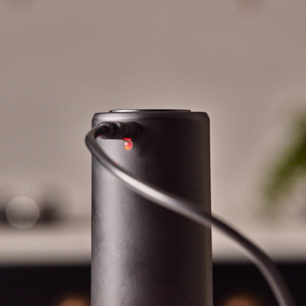 A Cole & Mason Brixton Electric Mill Matt Black GS, featuring a sleek cylindrical design with a power cord plugged in and a small red indicator light glowing near the port, is showcased against a blurred background that accentuates its convenient one-hand operation in the foreground.