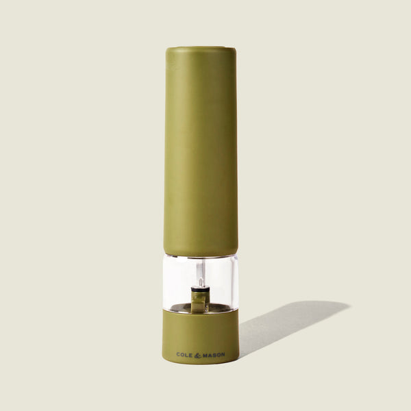 The Brixton Electric Mill Olive Green GS by Cole & Mason stands upright on a light beige background, displaying its olive green body, ceramic grinder, and transparent midsection with “COLE & MASON” written on the base, while casting a faint shadow.