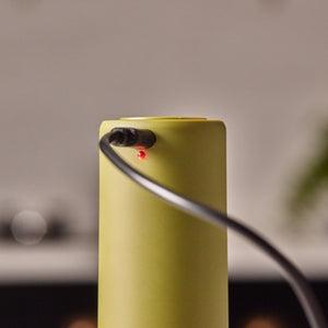 A close-up of the Cole & Mason Brixton Electric Mill Olive Green GS, a cylindrical electronic device designed for one-handed use, with a black cable plugged in and a small red indicator light turned on. The electric salt and pepper mill features a durable ceramic grinder for efficient seasoning.
