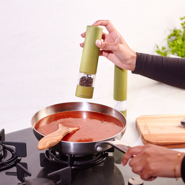 Using the Cole & Mason Brixton Electric Mill Olive Green GS with a durable ceramic grinder, someone effortlessly seasons a pan of red sauce simmering on the stovetop, operating the mill one-handed while a wooden spoon rests in the sauce and a cutting board sits in the background. This design makes seasoning quick and easy.