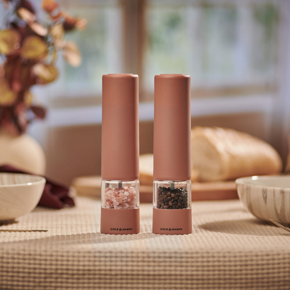 Two identical Cole & Mason Brixton Electric Mill Terracotta GS grinders stand side by side on a light beige background, casting soft shadows to the right. Featuring a durable ceramic grinding system and convenient USB-C charging, these mills combine modern functionality with elegant design.