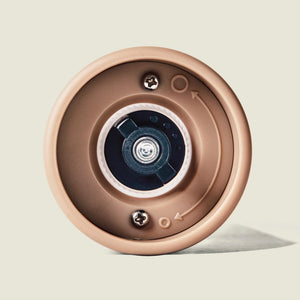 Close-up view of the bottom of the Cole & Mason Brixton Electric Mill Stone GS, a brown cylindrical electric mill. The metal mechanism features screws and directional arrows arranged in a circular pattern. The design includes an adjustable grind setting and is equipped with a durable ceramic grinding core for efficient performance.
