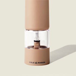 A tan Cole & Mason Brixton Electric Mill Stone GS pepper grinder with a clear middle section, featuring an adjustable grind and a durable ceramic grinding core, shown against a light beige background.
