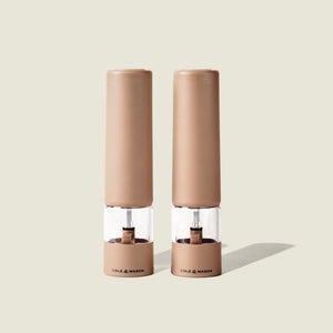 Two beige Cole & Mason Brixton Electric Mill Stone GS grinders with transparent midsections and the brand name printed at their bases, featuring adjustable grind settings and durable ceramic grinding cores, stand side by side on a light neutral background.
