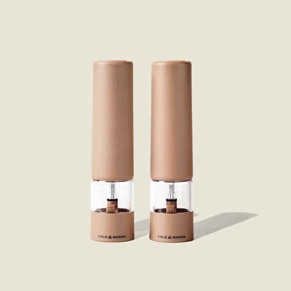 Two beige Cole & Mason Brixton Electric Mill Stone GS grinders with transparent midsections and the brand name printed at their bases, featuring adjustable grind settings and durable ceramic grinding cores, stand side by side on a light neutral background.