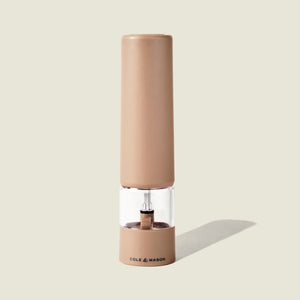 A tan-colored Brixton Electric Mill Stone GS pepper grinder by Cole & Mason stands upright on a light beige background, casting a shadow to the right. It features a transparent middle section, an adjustable grind setting, and a durable ceramic grinding core. The Cole & Mason brand is printed on the bottom front.