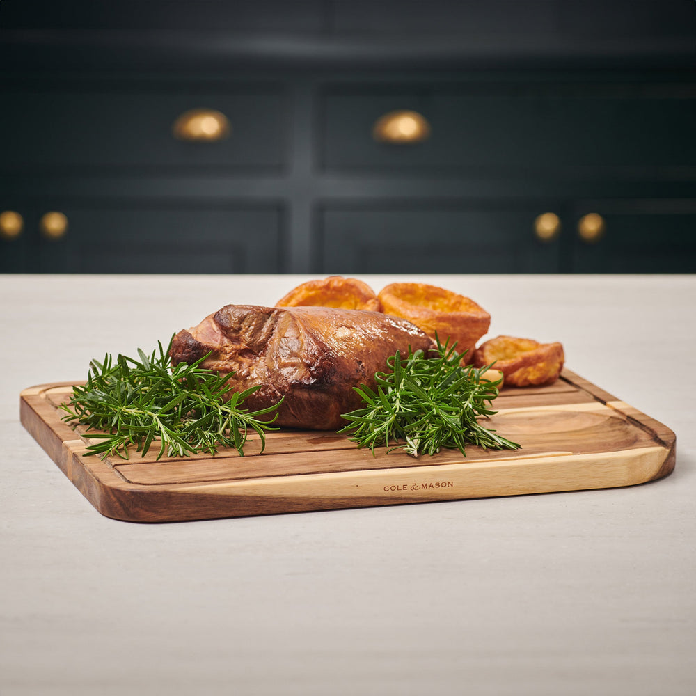 The Cole & Mason Berden Acacia Carving Board features a rectangular design with rounded corners, a recessed juice groove, and a circular indentation. Its striped acacia wood pattern adds style to your kitchen.