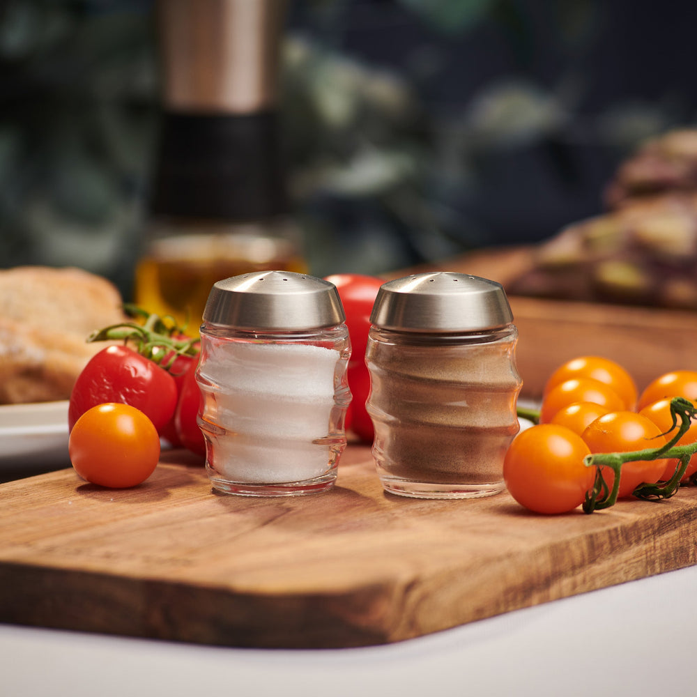 The Cole & Mason Bray Glass Shakers Set features two clear glass shakers with stainless steel tops and a wavy design—one for salt, one for pepper—placed side by side, ideal for any kitchen décor.