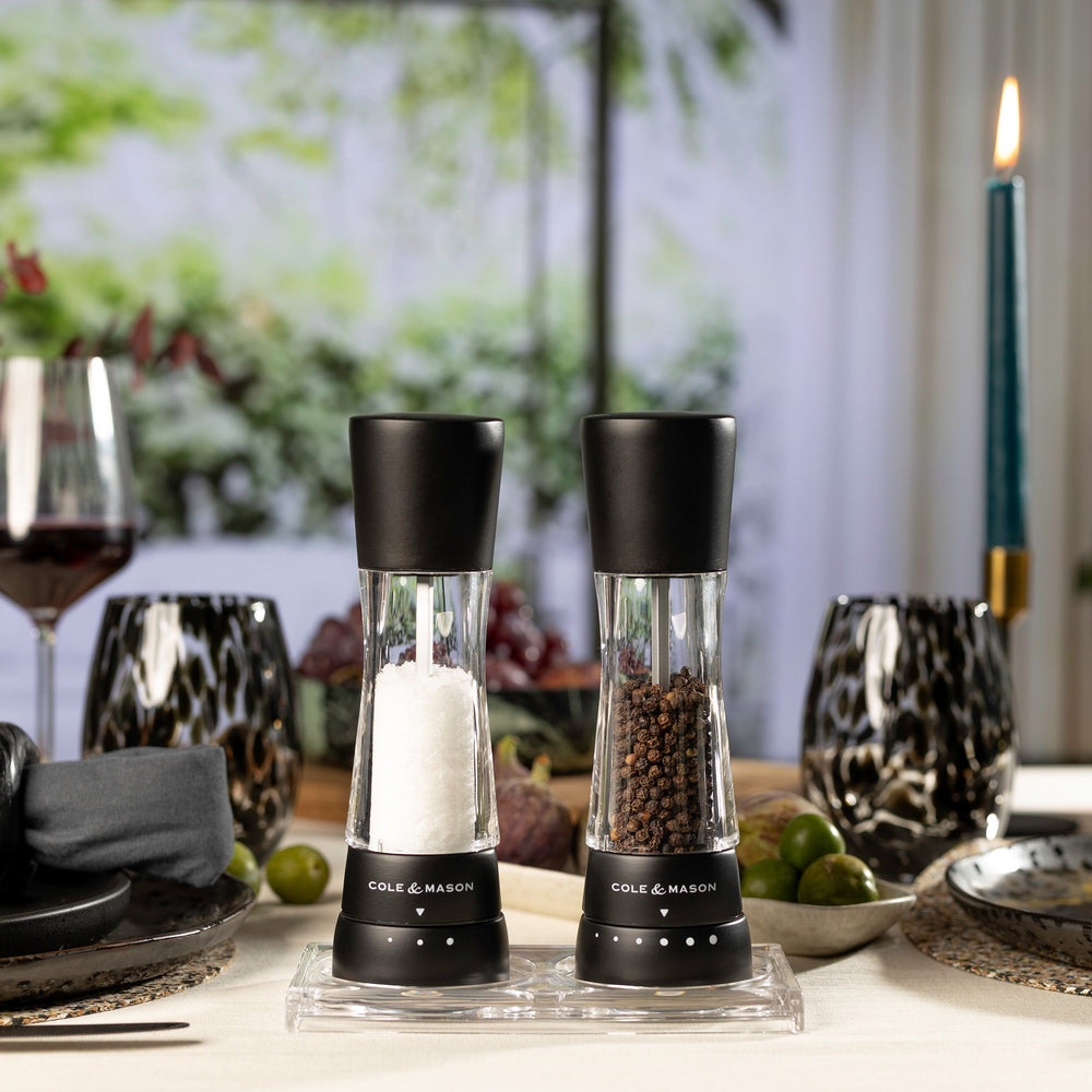 #salt&pepperset Two Cole & Mason Derwent Matt Black Salt & Pepper Mills, featuring adjustable grind settings and acrylic bodies, stand together on a light background, filled with coarse salt and whole black peppercorns.