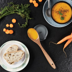 A bowl of carrot soup with herbs, a wooden spoon holding soup on the Cole & Mason Duxford Ceramic Spoon Rest, sliced bread, and fresh carrots with greens are artfully arranged—an ideal scene for elegant kitchen decor.