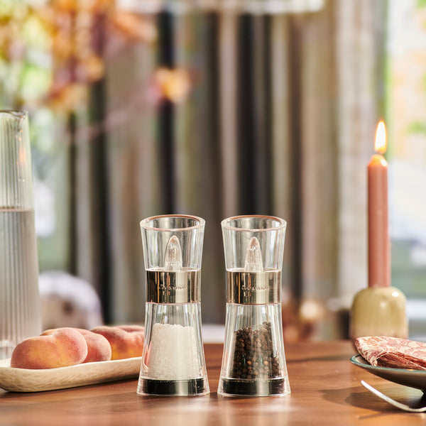 The Cole & Mason Flip Salt & Pepper Mill Set, filled with Belgian sea salt and Vietnamese peppercorns, sits on a wooden table with peaches, napkins on a plate, a lit candle, and a glass jug of water for an inviting touch.