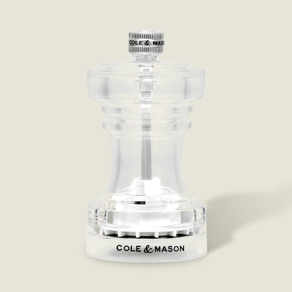 Showcased against a light beige background, the Cole & Mason Acrylic Salt Shaker & Pepper Mill Set (104 mm) features a clear acrylic body with a silver knob on top and “COLE & MASON” inscribed in black on the base; this elegant pepper mill offers an adjustable grinder for customized seasoning.