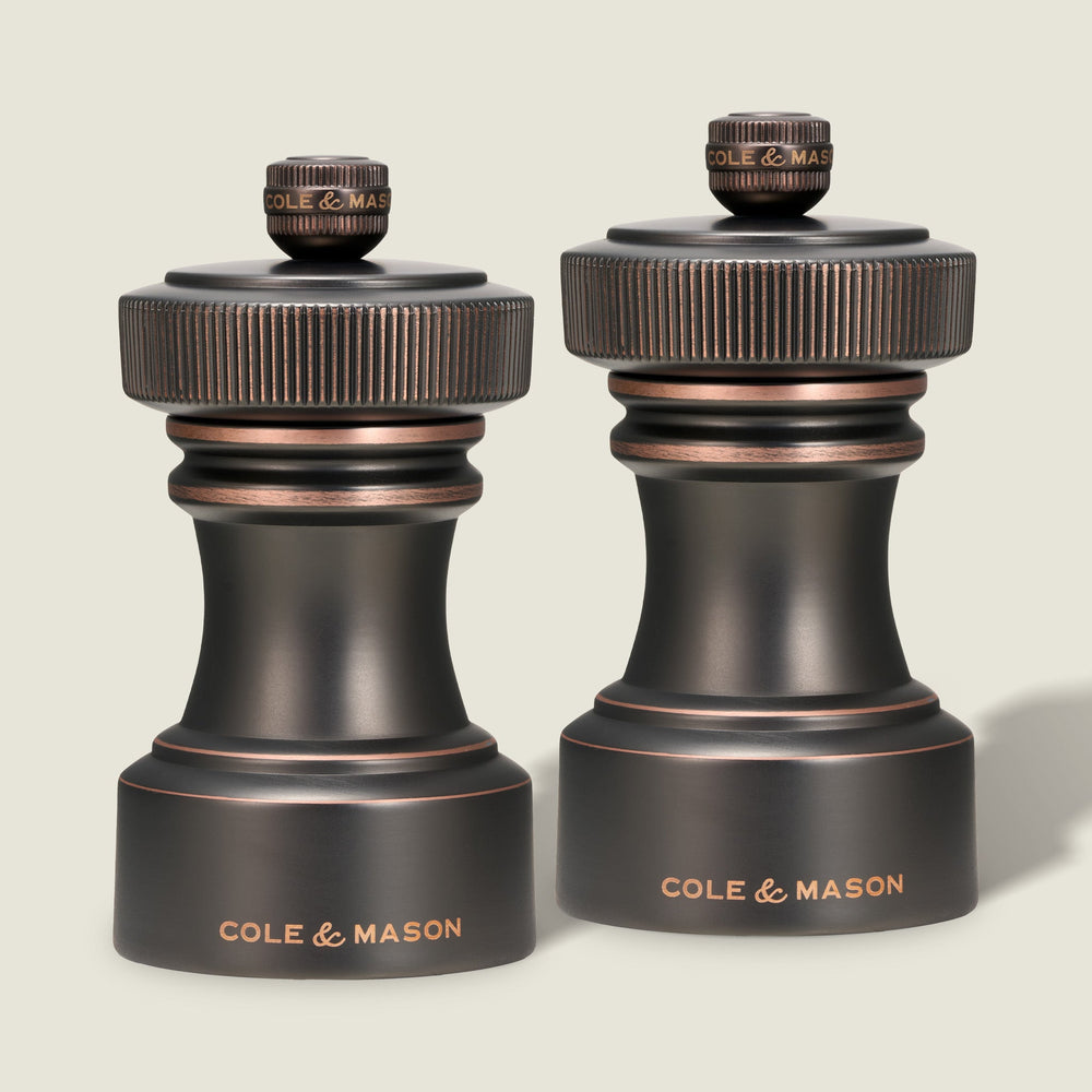 Two Cole & Mason Hoxton Salt and Pepper Mills in antique bronze, each with a dark finish, textured knurled tops, and the Cole & Mason brand name engraved in gold at the base, stand side by side against a plain light background. This sophisticated set features a precision grinding system for effortless seasoning every time.