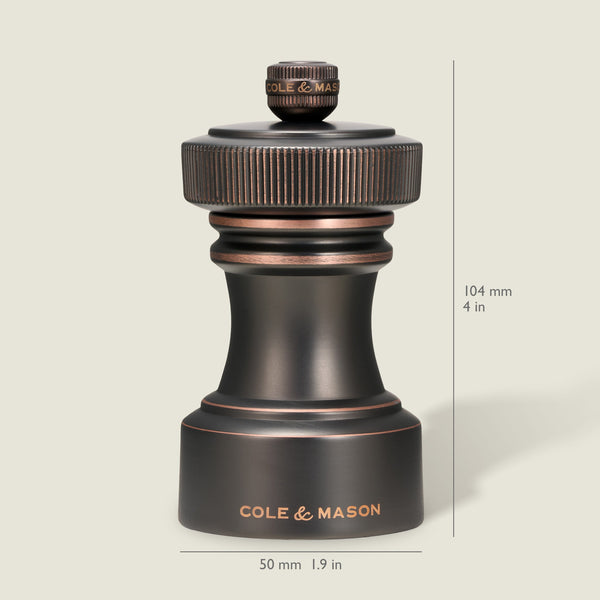A dark metallic pepper grinder from the Cole & Mason Hoxton Salt and Pepper Mill Set in an antique bronze finish is shown upright against a light background, labeled with the brand name. The mill measures 104 mm (4 in) tall and 50 mm (1.9 in) wide, and features a precise grinding system for consistent seasoning—perfect for pairing with any salt or pepper mill set.