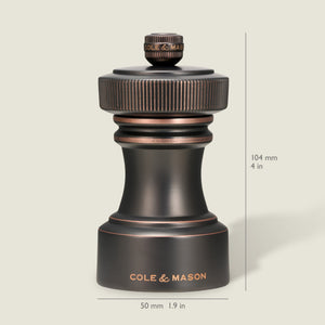A dark metallic pepper grinder from the Cole & Mason Hoxton Salt and Pepper Mill Set stands upright, showcasing an antique bronze finish. Dimensions are indicated as 104 mm (4 in) tall and 50 mm (1.9 in) wide, set against a light beige background.