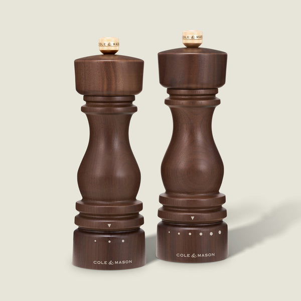 Two Cole & Mason London Walnut Salt or Pepper Mills stand side by side on a neutral background. The pepper mill is positioned on the left, while the adjustable salt grinder sits on the right; each features distinct grind size indicators at the base.