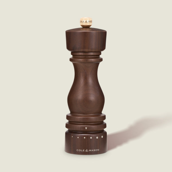 The Cole & Mason London Walnut Salt or Pepper Mill, featuring a rounded dark wood walnut design and labeled with the brand name at the base, stands upright on a light beige background. Adjustment dots and a small knob are visible at the top and base of this elegant seasoning mill.