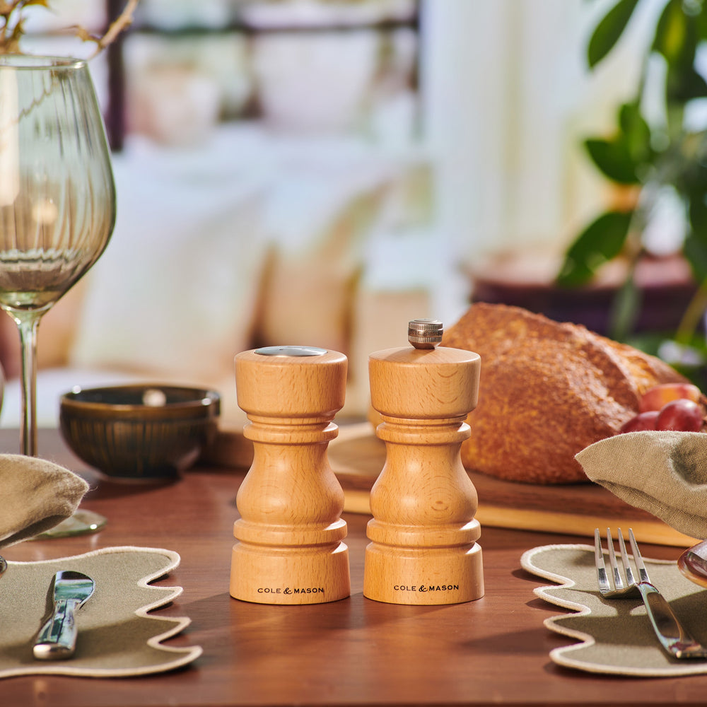 The Cole & Mason London Natural Beech Salt Shaker & Pepper Mill Set, crafted from natural beech wood, stands upright on a light surface. The salt shaker features a silver knob with COLE & MASON engraved, reflecting their elegant design.