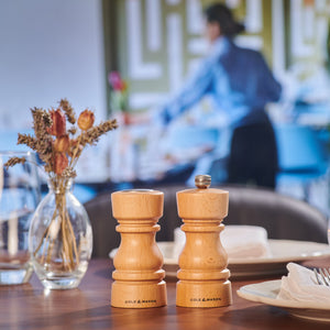 A Cole & Mason London Natural Beech Salt Shaker & Pepper Mill Set rests on a restaurant table with plates, napkins, and dried flowers in a glass vase, while a blurred figure works in the background.