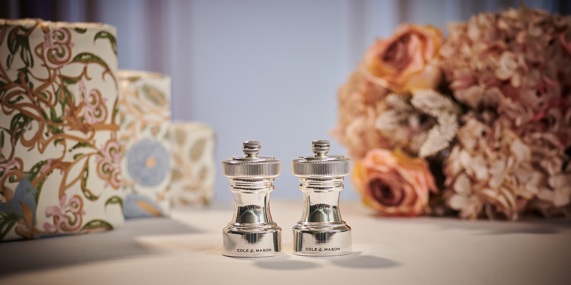 Two silver salt and pepper grinders are placed side by side on a table, with floral-patterned boxes and a bouquet of pink and beige flowers in the blurred background.