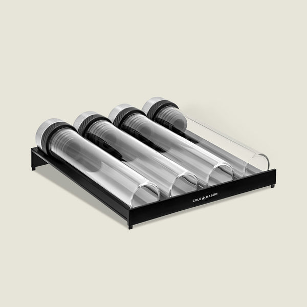 A set of four clear, airtight glass jars with black lids, laid horizontally in a sleek black rack labeled Cole & Mason—a stylish Shadwell Drawer-Fit Spice Storage set for your kitchen.