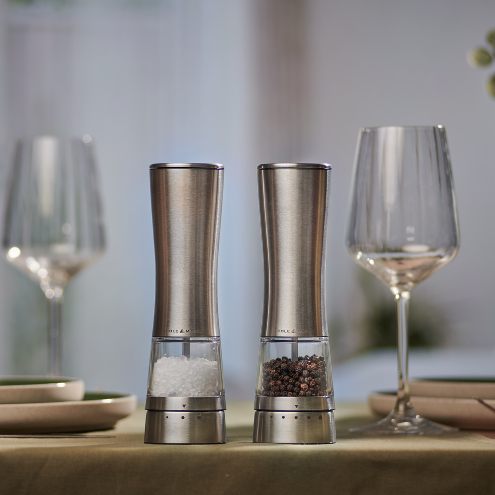 Two Cole & Mason Electric Derwent Salt & Pepper Mills stand side by side; one filled with coarse salt, the other with whole black peppercorns. Both feature stainless steel construction, clear bottoms, and adjustable grind settings.